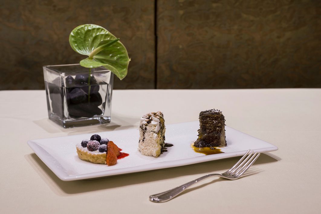 На белой прямоугольной тарелке лежат три небольших десерта, рядом — стеклянная ваза с одним листочком и вилкой.