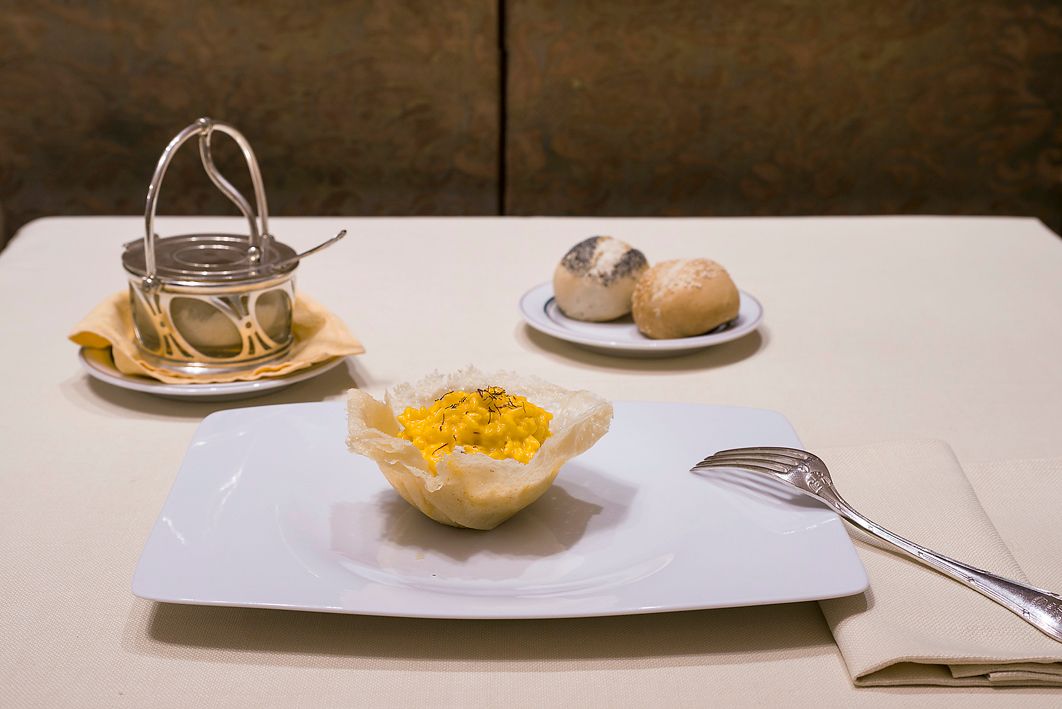 Миска желтого риса в хрустящей слоеной чашечке, подается на белой тарелке с хлебом, баночкой с приправами и серебряной ложкой.