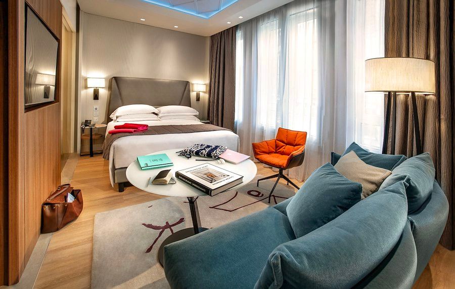 酒店套房内设有一张特大号床、一张蓝色沙发、一把橙色扶手椅和一张圆形桌子，铺着一块花纹地毯。