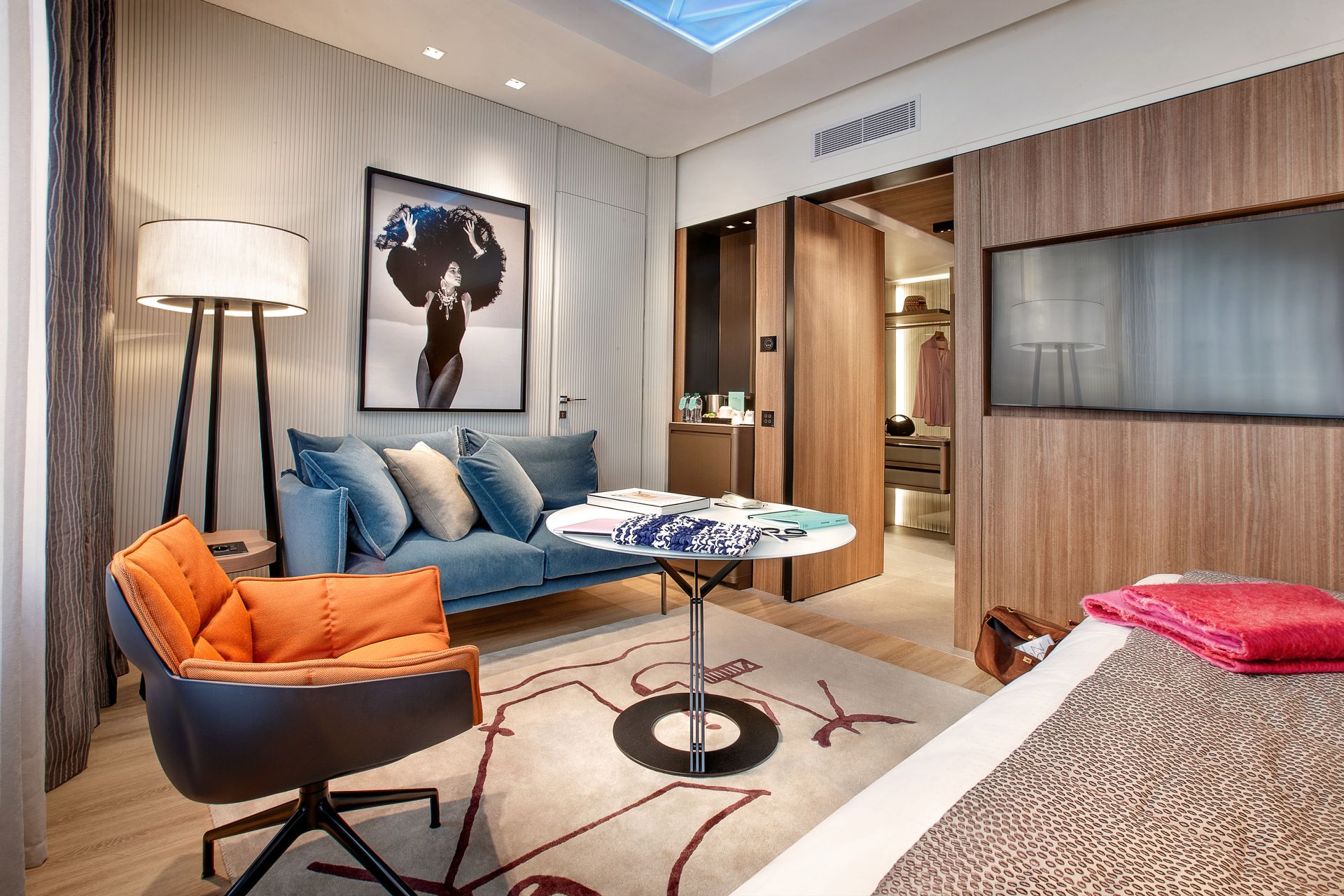 一间现代化的酒店房间，配有橙色椅子、蓝色沙发、小餐桌、墙面艺术品和步入式衣橱。