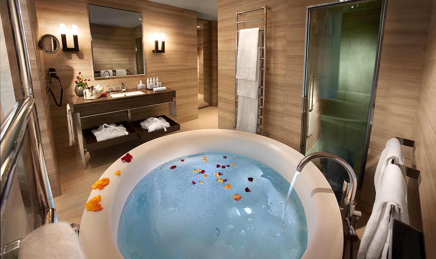 A round bathtub filled with blue water and flower petals, set in a modern, neutral-toned hotel bathroom.
