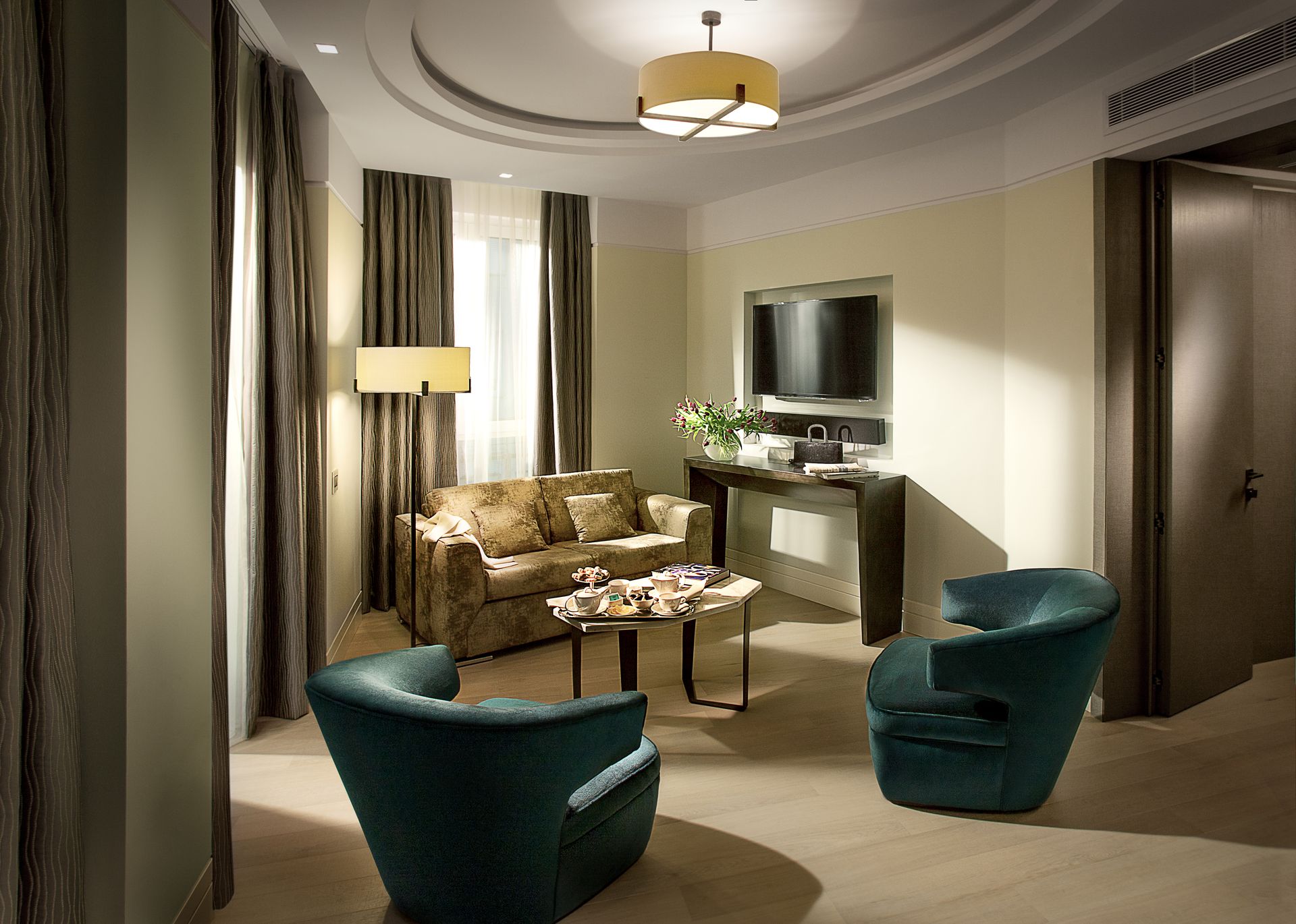 A luxury hotel suite featuring a beige sofa, two teal velvet armchairs, a round coffee table, and a wall-mounted TV.