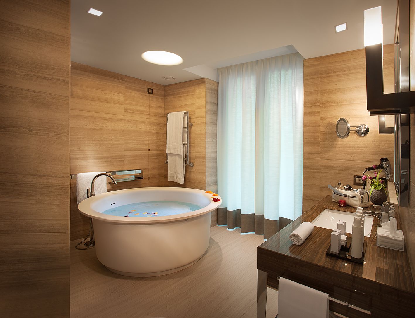 Светлая, современная ванная комната с деревянными панелями на стенах, большой овальной ванной и туалетным столиком у окна со шторами.