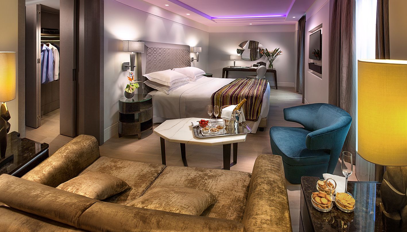 Роскошный гостиничный номер с мягким диваном бежевого цвета, белым журнальным столиком с напитками, большой кроватью и синим креслом.