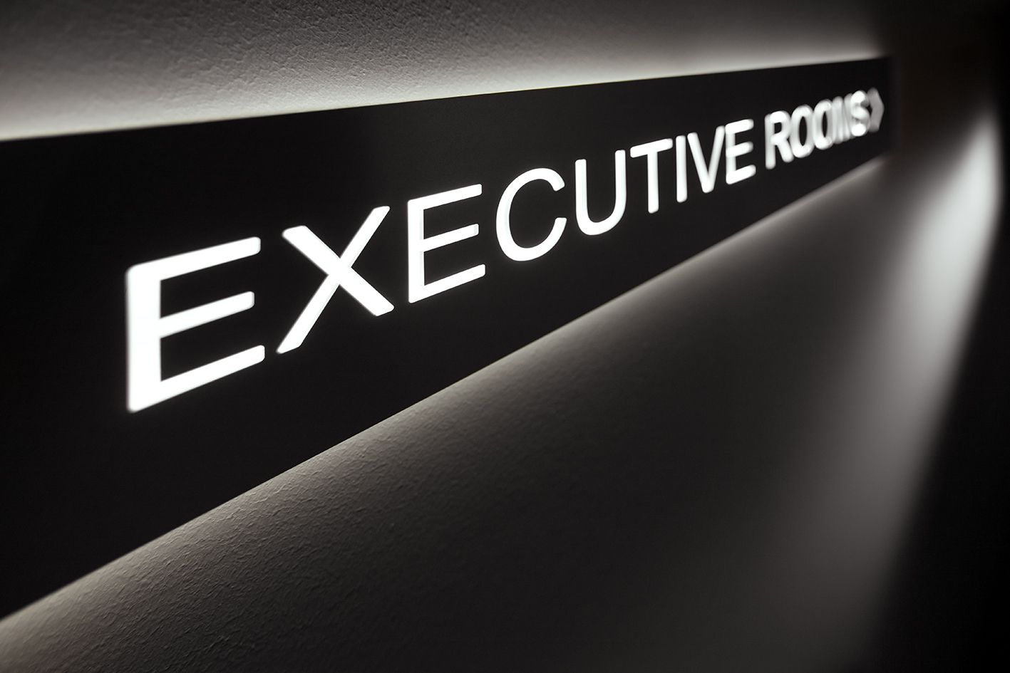 На темной стене с подсветкой висит надпись «EXECUTIVE ROOMS» (Комнаты для руководителей), выполненная жирными белыми заглавными буквами и указывающая вправо стрелкой.