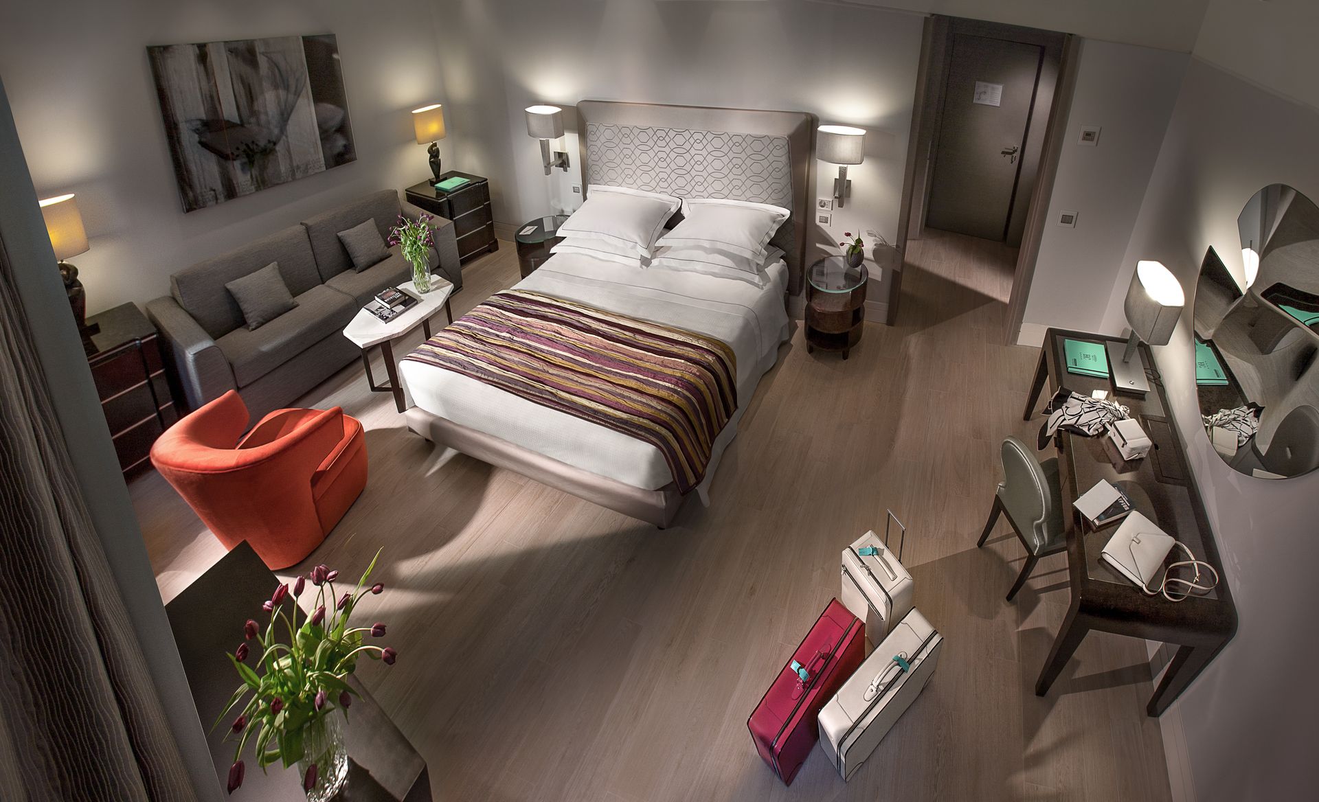 Вид сверху на гостиничный номер, в котором находятся двуспальная кровать, серый диван, оранжевое кресло, письменный стол и два чемодана на полу.