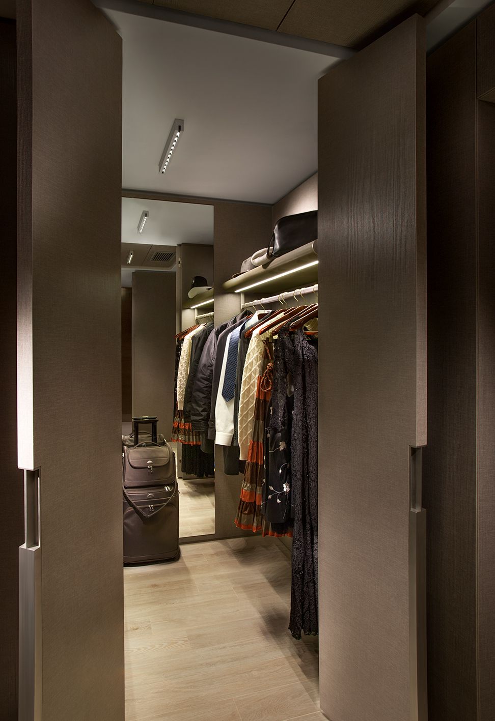 Гардеробная комната с шкафами бежевого цвета, развешенной одеждой, полкой с подсветкой и чемоданом на полу.