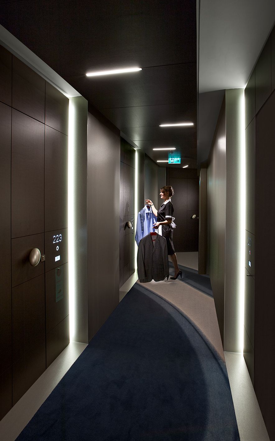 Человек стоит в современном, выкрашенном в темные тона коридоре отеля, держа в руках костюм на вешалке возле дверного проема.