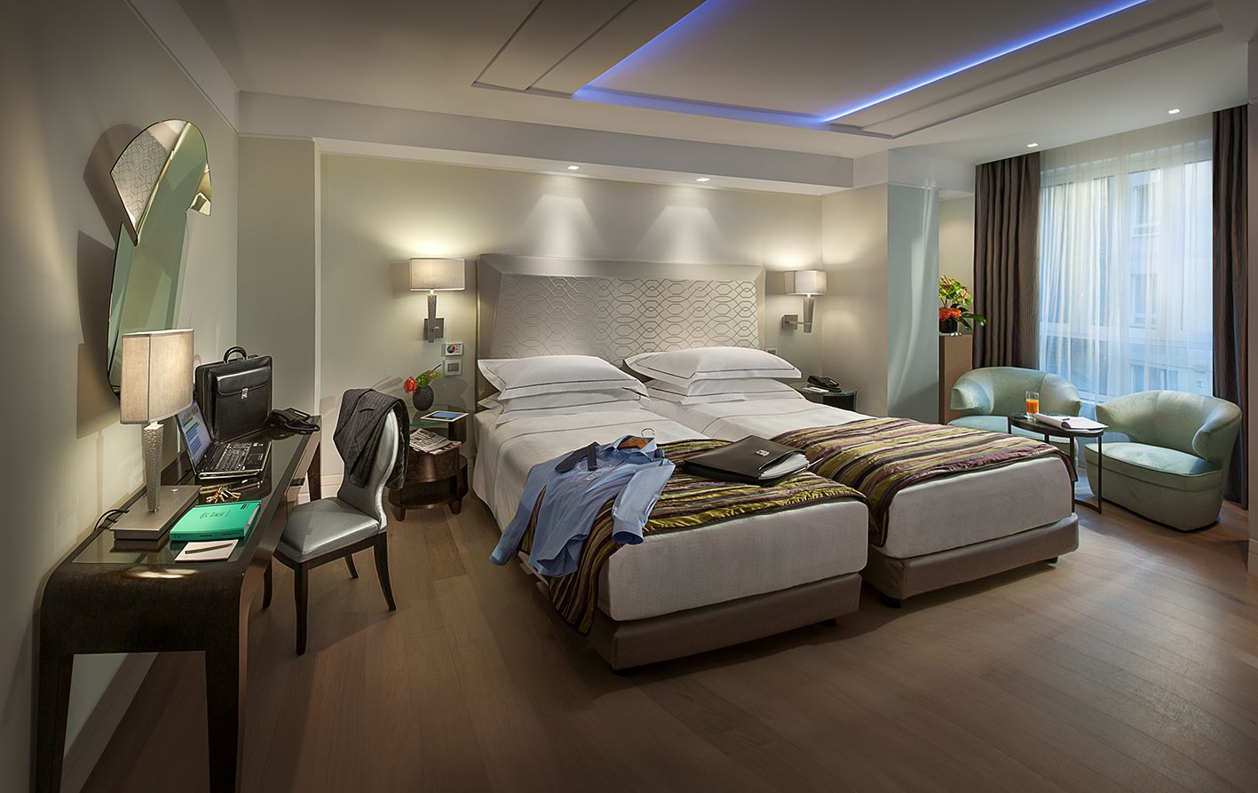 Современный гостиничный номер с двумя односпальными кроватями, письменным столом, стулом и двумя креслами у окна с полупрозрачными занавесками.