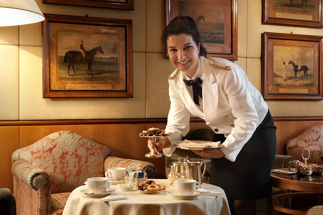 Официант в белой униформе улыбается, ставя многоярусный поднос с выпечкой на сервированный стол в элегантной чайной комнате.