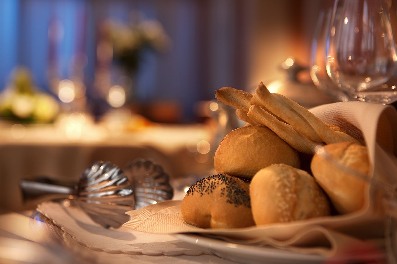 Корзина с разнообразными булочками и хлебными палочками, поданными на тканевой салфетке, на фоне размытого изображения ресторанной обстановки.
