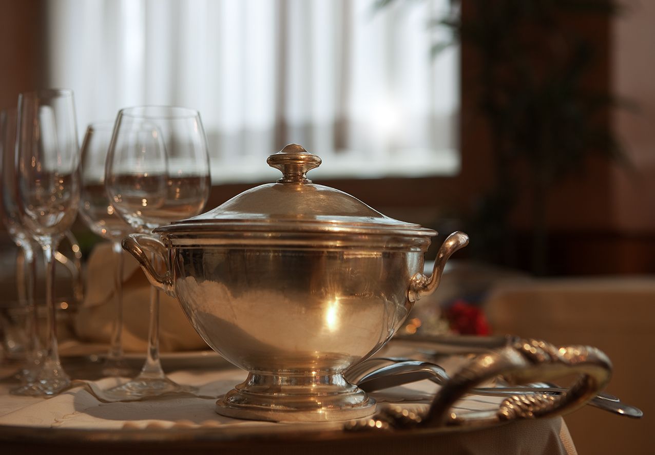 В мягко освещенной комнате на белой скатерти стоит серебряное блюдо с крышкой, а на столе — бокалы для вина.
