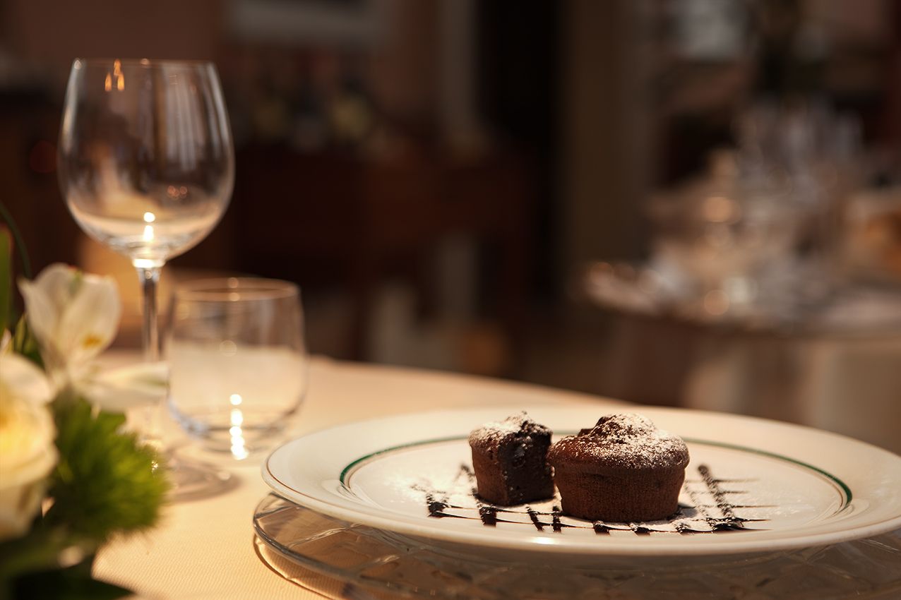 Два шоколадных маффина, посыпанных сахарной пудрой, лежат на тарелке, на обеденном столе в окружении бокалов и цветов.
