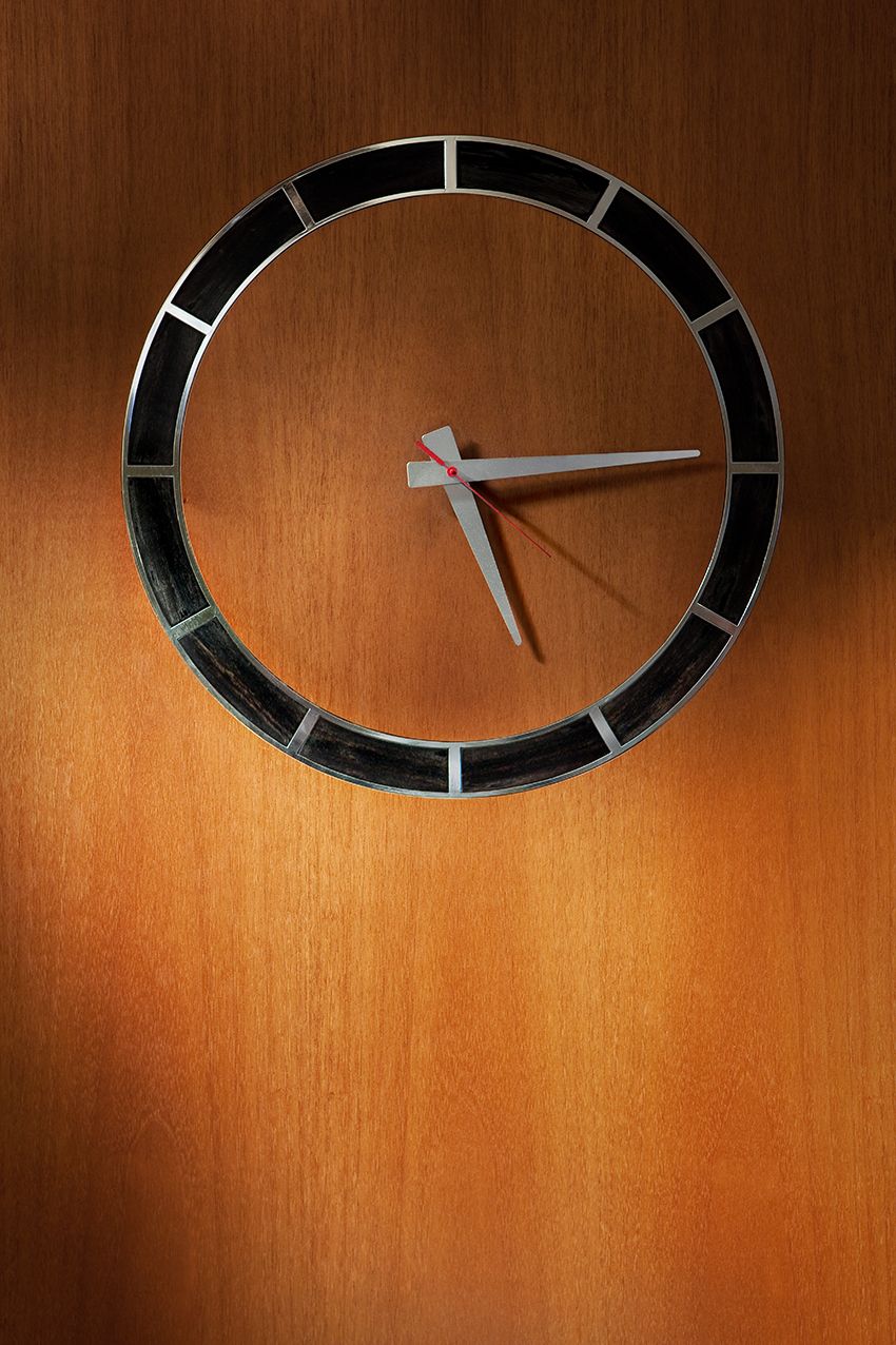 Минималистичные часы с черной круглой рамкой и светлыми стрелками, установленные на фактурной стене, отделанной теплыми деревянными панелями.