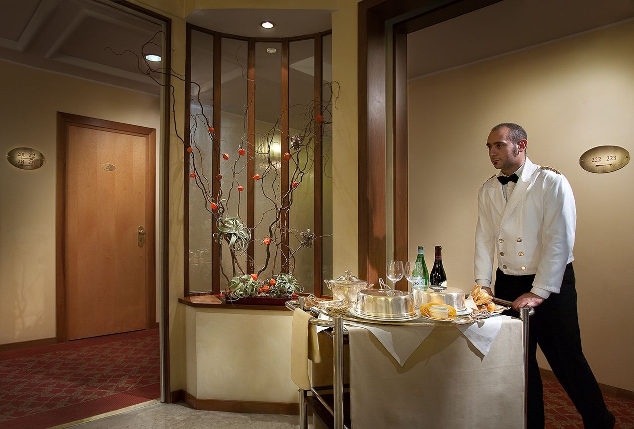 Официант в белой униформе катит по коридору отеля серебристую тележку для обслуживания номеров, между двумя дверями которой находится цветочная композиция.