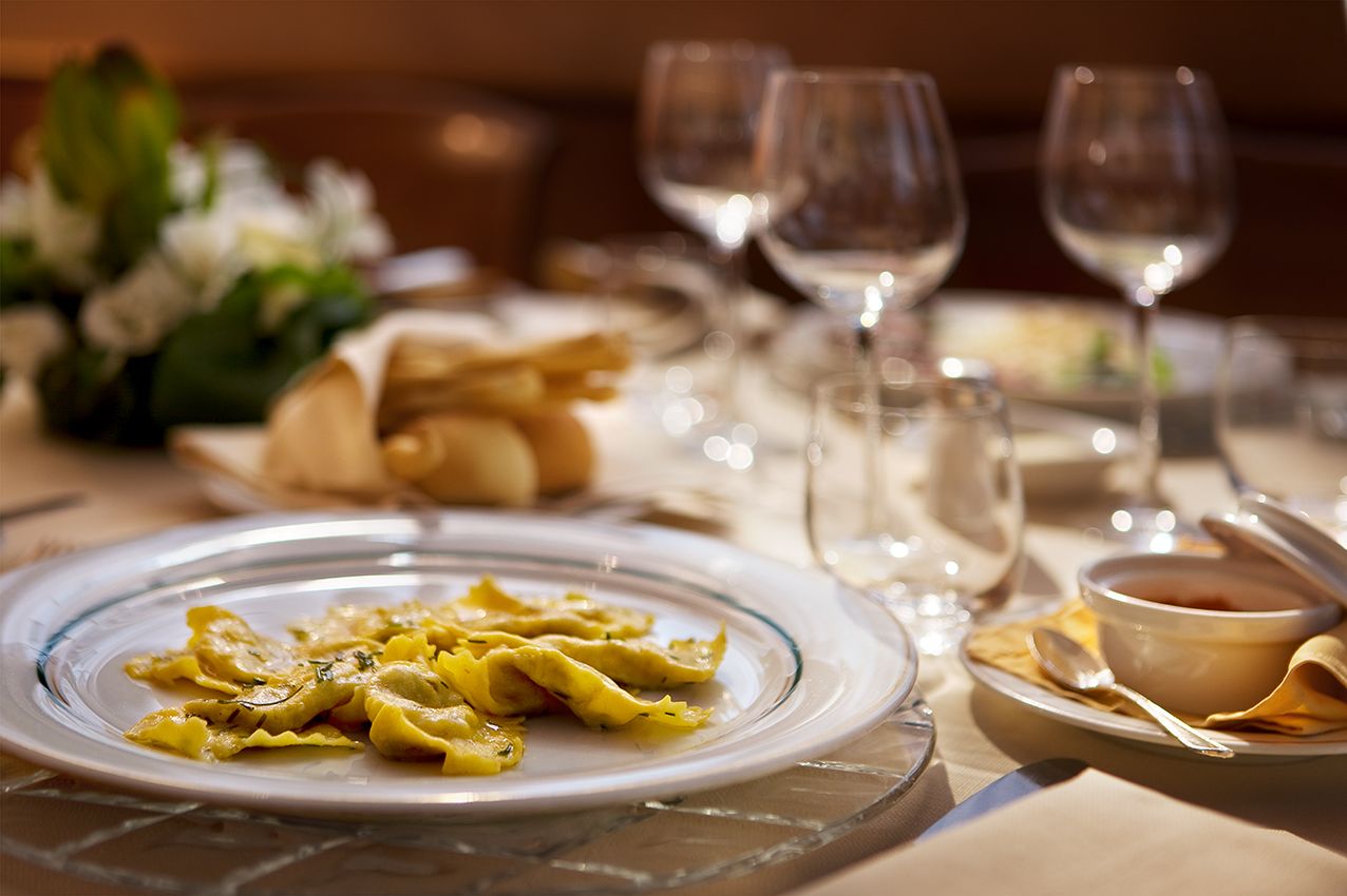 На сервированном обеденном столе стоит белая тарелка с золотистыми равиоли, а также бокалы для вина, хлебные палочки и небольшая мисочка с соусом.