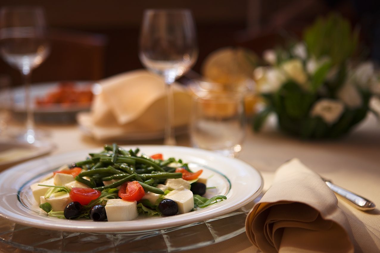 На обеденном столе тарелка салата из зеленой фасоли, моцареллы, помидоров и оливок, а также стаканы и салфетка.
