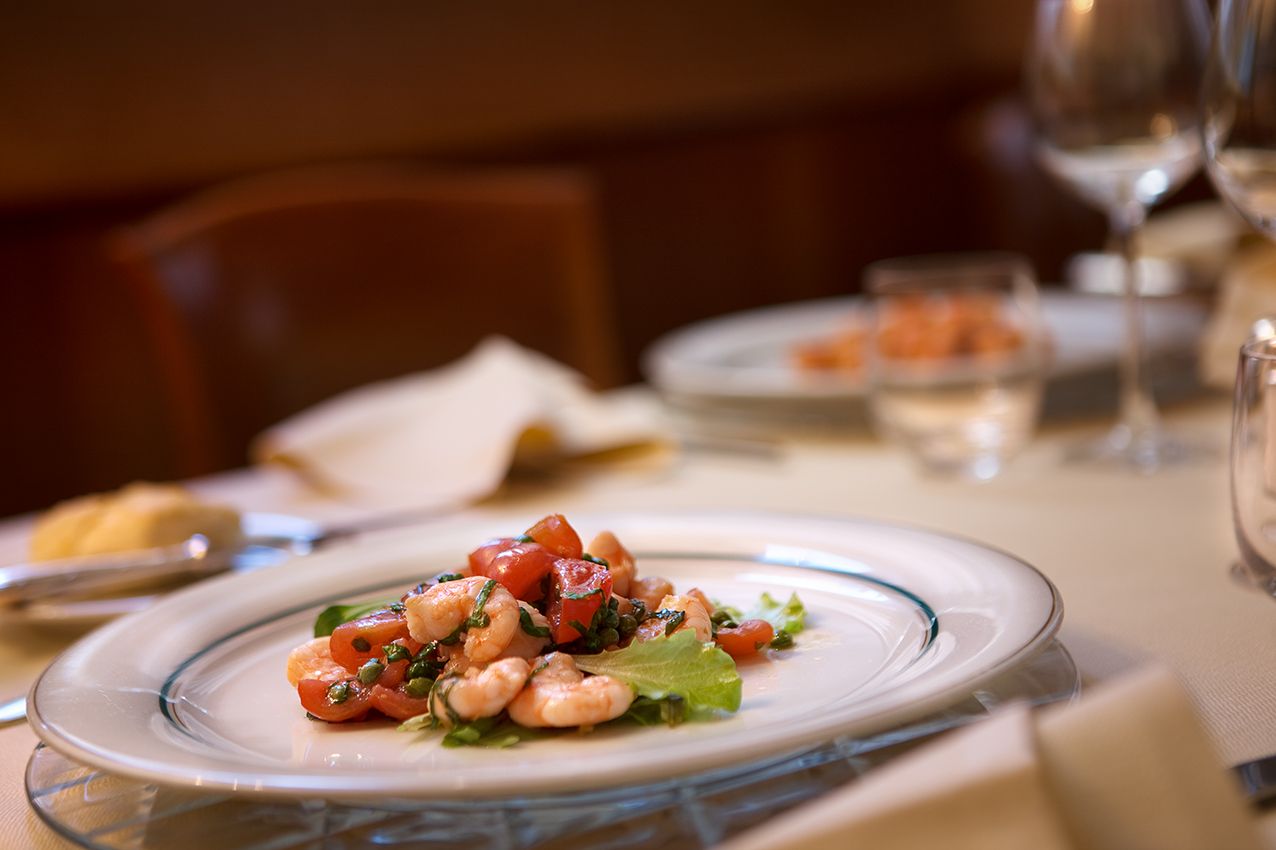 一道虾仁番茄开胃菜盛放在白色盘子里，摆放在餐厅的餐桌上，背景中可以看到酒杯。