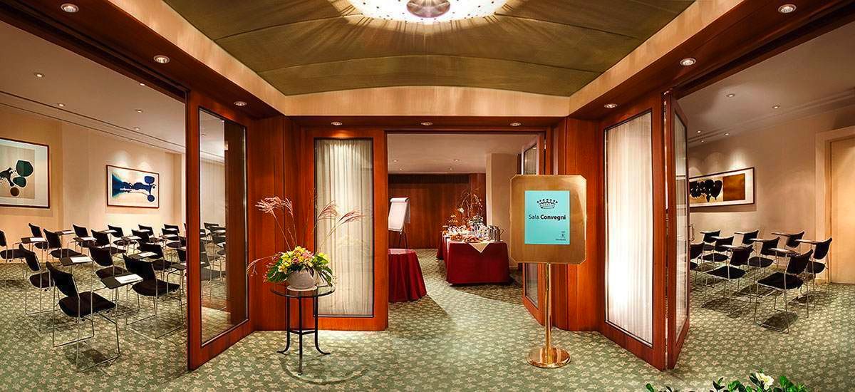A formal hotel meeting room with light-colored walls, patterned carpeting, wooden partitions, and rows of seating.