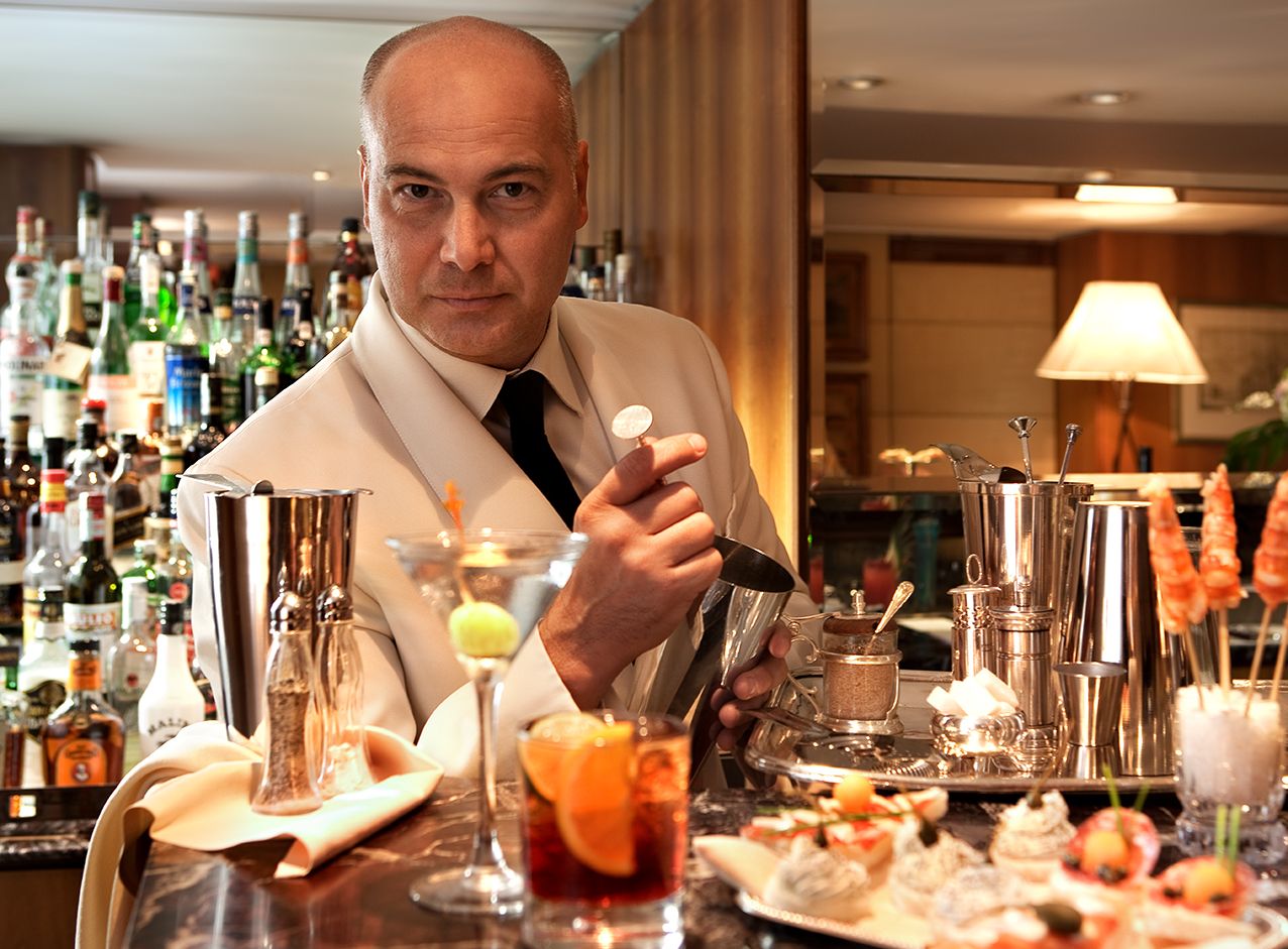 Бармен в белом пиджаке готовит напитки за хорошо укомплектованной барной стойкой, где представлены различные коктейли и закуски.