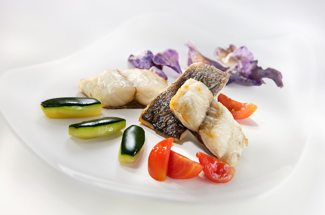 Изысканное блюдо, подаваемое порционно, включает в себя обжаренное филе белой рыбы, дольки цуккини, ломтики помидоров и съедобные цветы фиолетового цвета.
