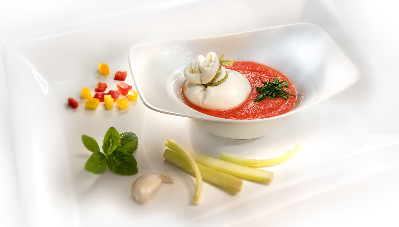 Порция сыра буррата, подаваемая в красном соусе, украшенная зеленью и сервированная перцем, мятой и ароматными травами.