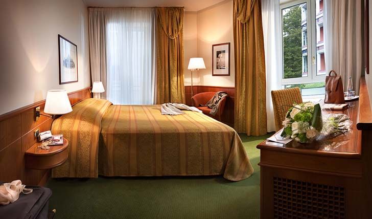 Уютный гостиничный номер с аккуратно заправленной кроватью, горчичными занавесками, деревянным столом и букетом цветов у окна.