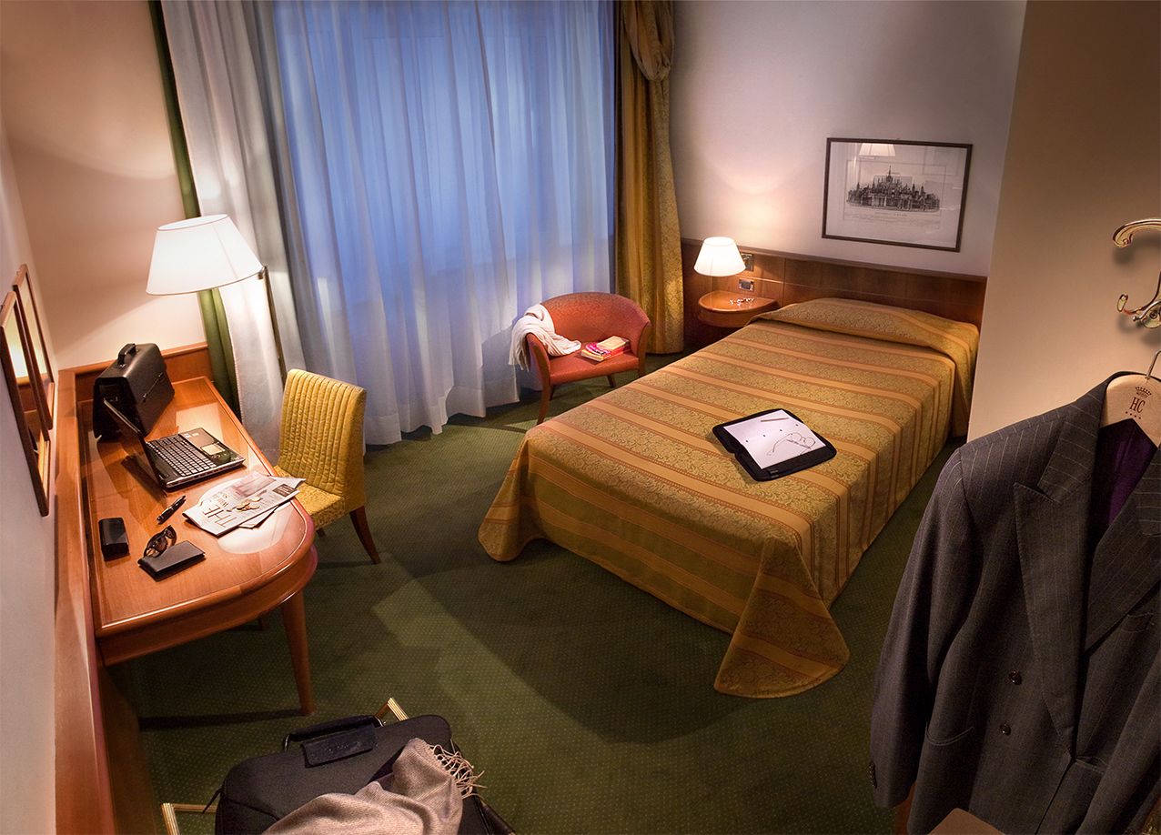 Уютный гостиничный номер с односпальной кроватью, деревянным столом с ноутбуком, зеленым ковровым покрытием и стулом у окна.