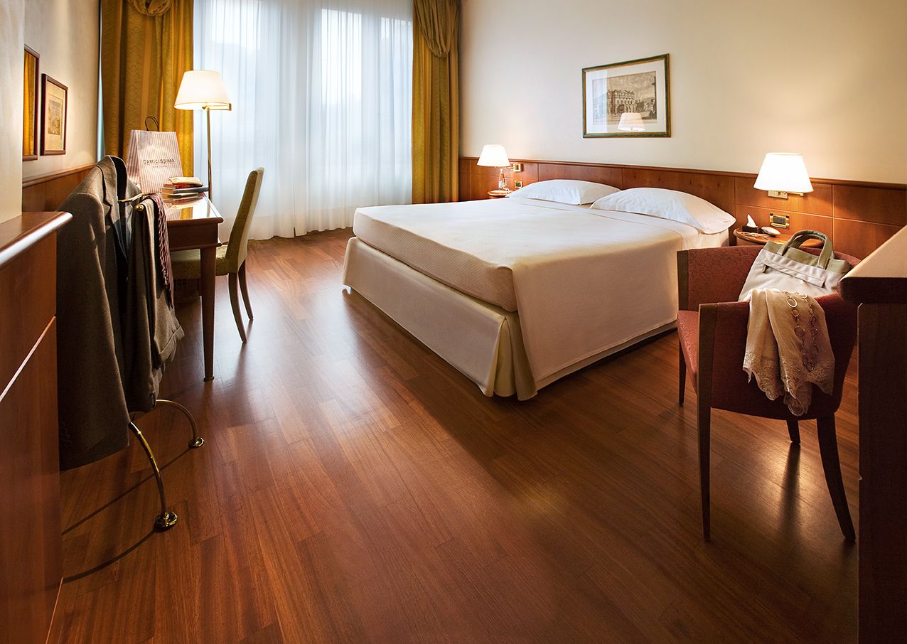 Гостиничный номер с аккуратно застеленной двуспальной кроватью, деревянным полом, письменным столом с лампой, стулом и вешалкой для одежды.