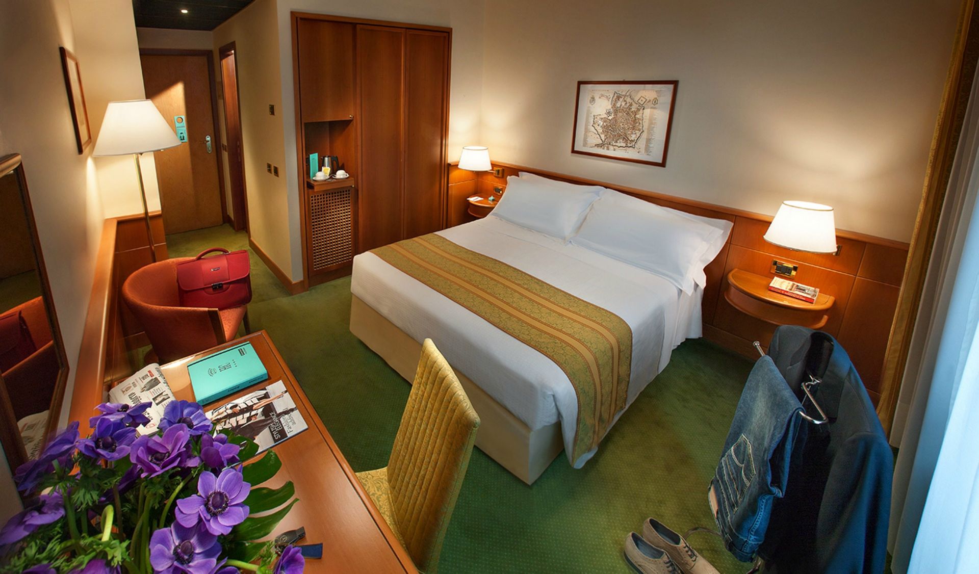 酒店房间内有一张大床、一张木桌和一个衣柜，前景是紫色的花朵，铺着绿色的地毯。