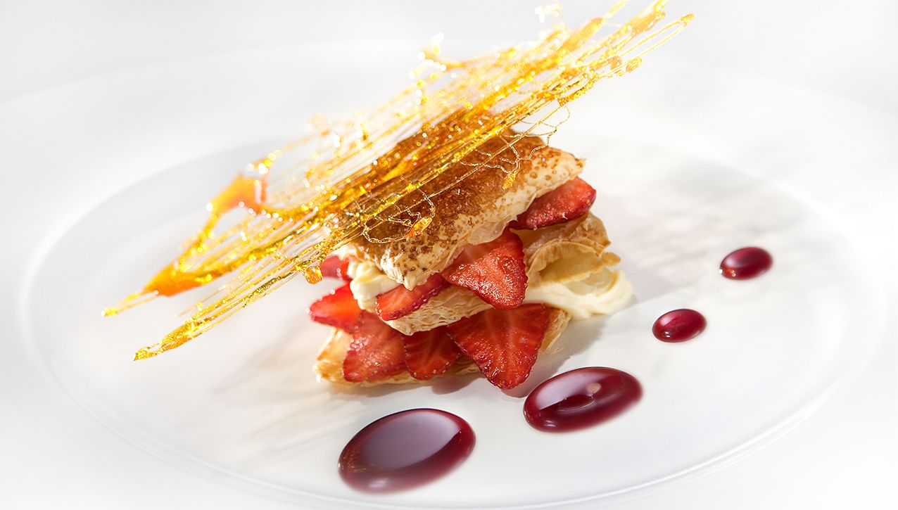 Клубничный мильфей, прослоенный кремом, посыпанный сахарной нитью и подаваемый с фруктовым соусом на белой тарелке.