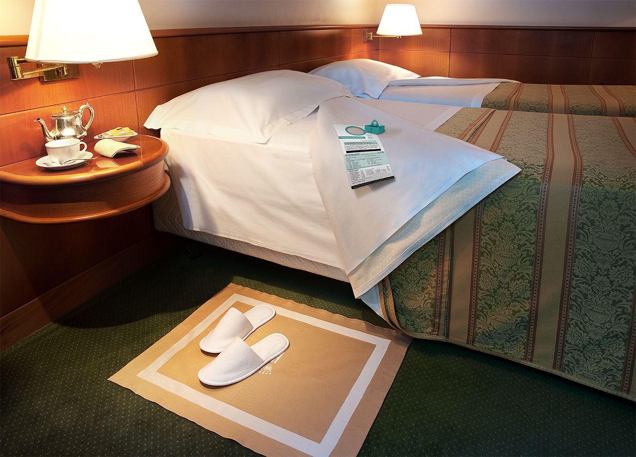 В гостиничном номере две односпальные кровати, деревянное изголовье, лампы, прикроватный столик и тапочки, лежащие на ковре у кровати.