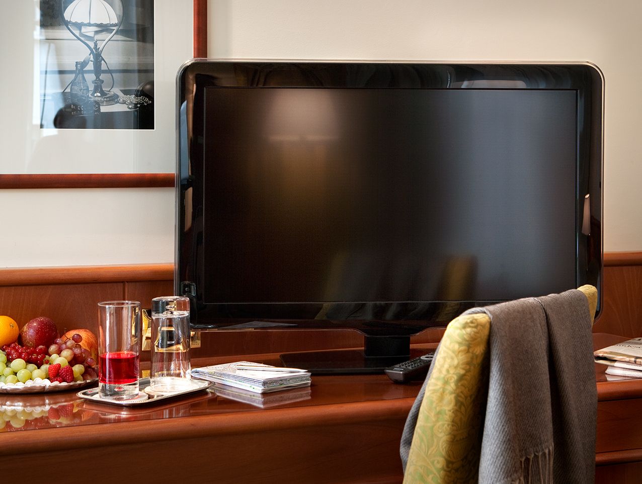 В номере, оформленном в гостиничном стиле, на деревянном столе стоит телевизор, а рядом — миска с фруктами, два стакана и стул, покрытый драпировкой.