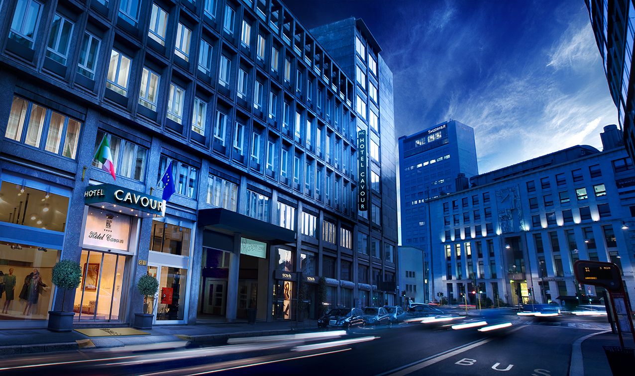 City street scene at dusk with a large hotel building, storefronts, and motion-blurred cars on a blue-toned road.