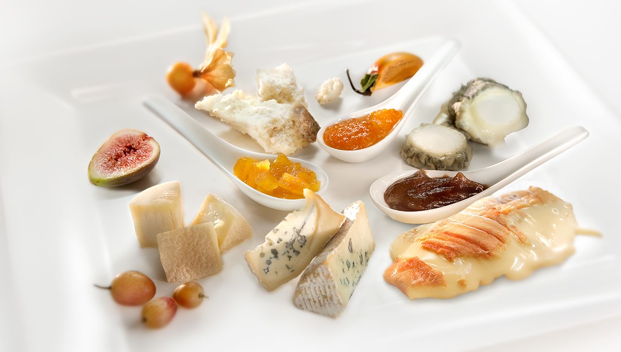 На белом блюде элегантно разложены разнообразные сыры ручной работы, свежие фрукты и небольшие ложечки джема.