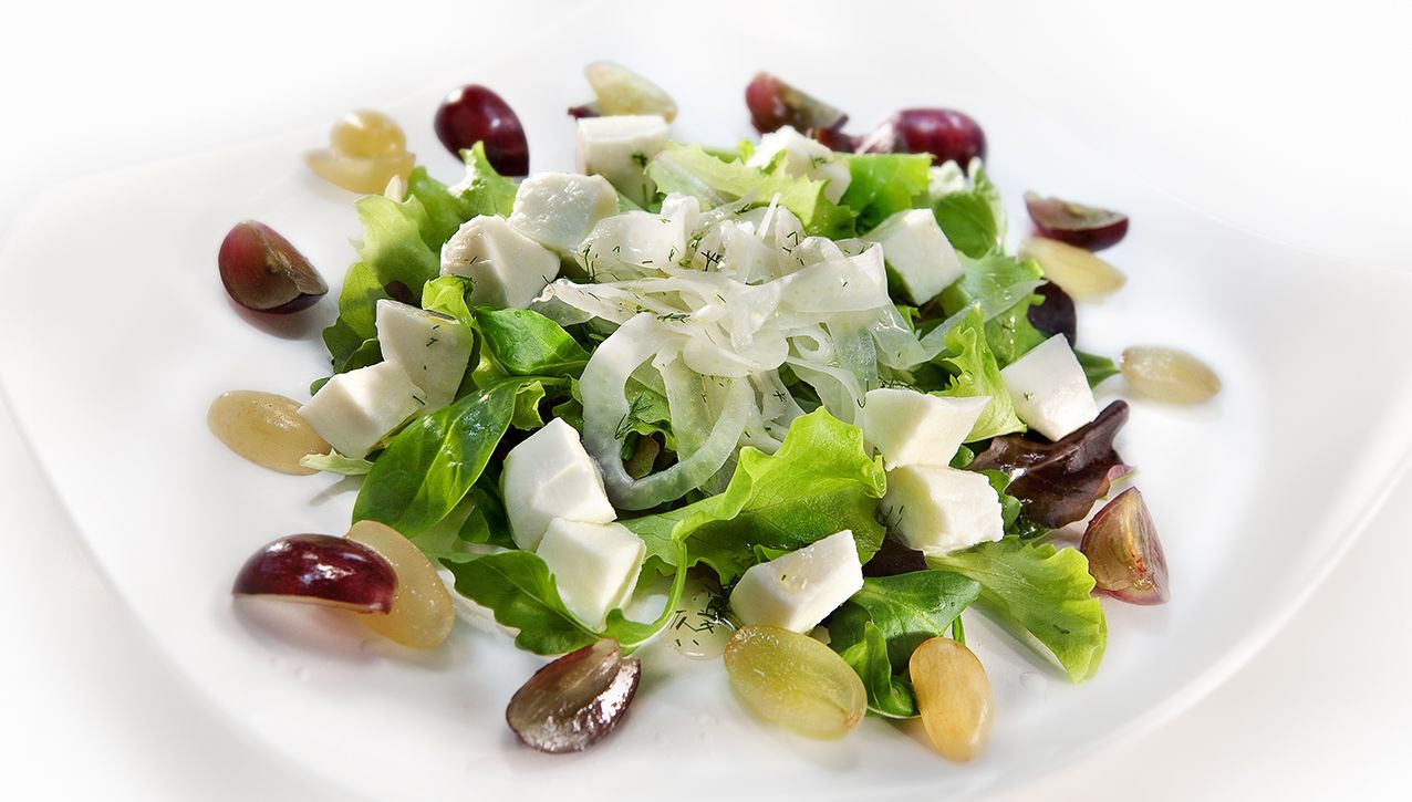 Салат из зелёного салата, кубиков белого сыра и нарезанного красного и зелёного винограда на белой тарелке.