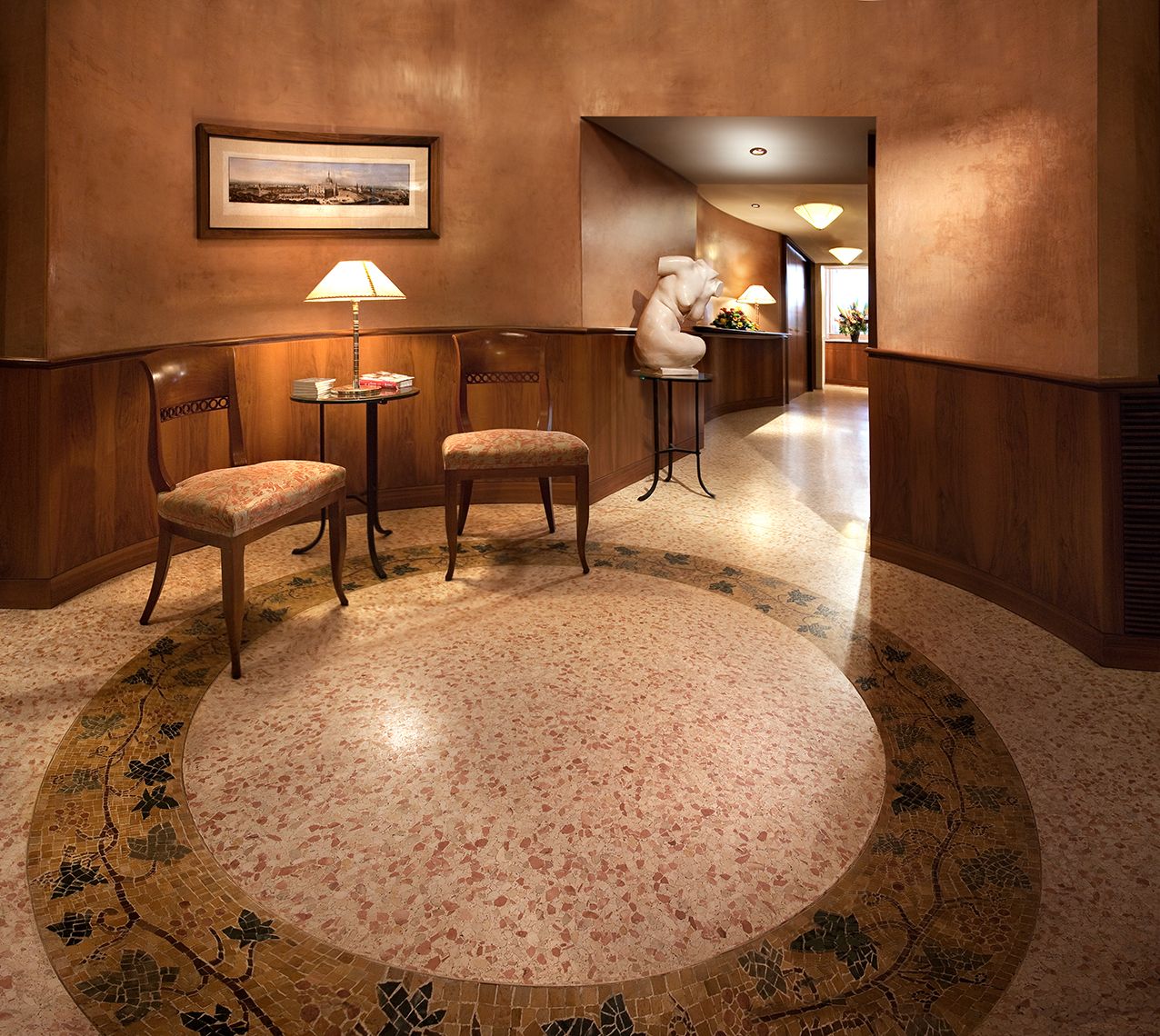 Небольшая зона отдыха с двумя деревянными стульями, приставным столиком с лампой и мраморным полом с круговым мозаичным узором.