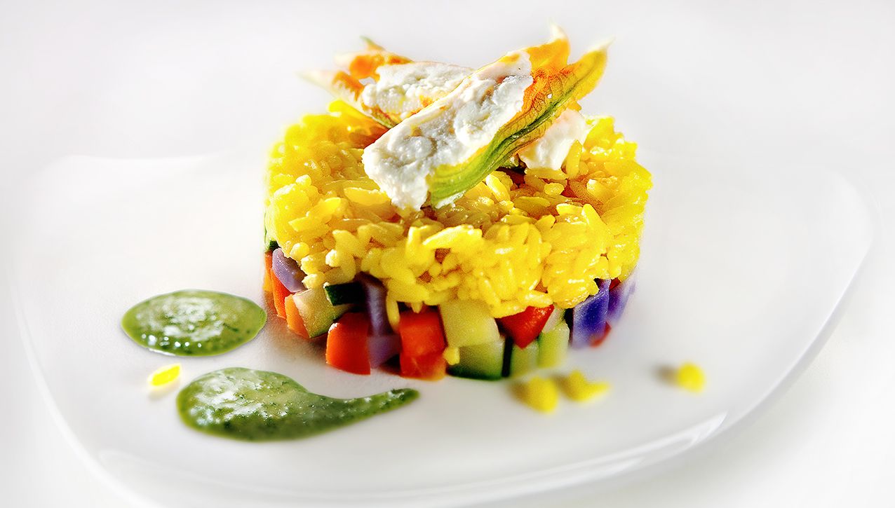 Красочная башня из овощей и желтого риса, увенчанная цветком цуккини, подается с зеленым соусом на белой тарелке.
