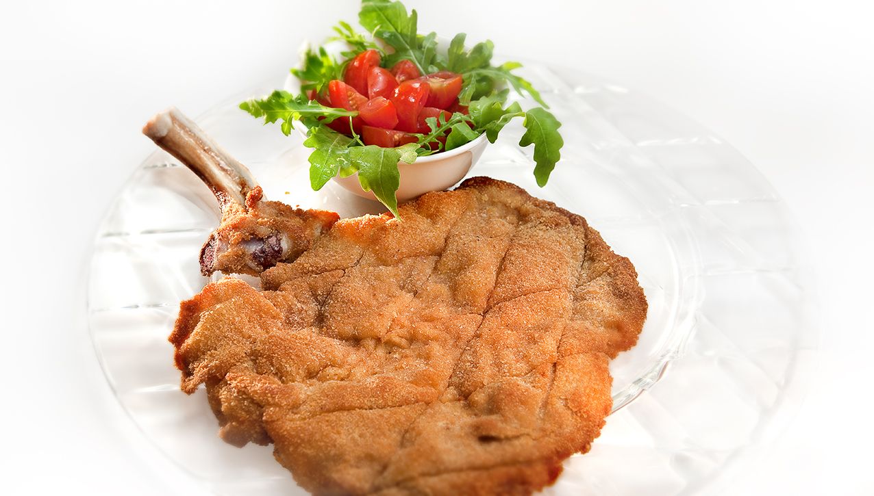 Панированная свиная отбивная на кости, подается на стеклянной тарелке с небольшим гарниром из салата из рукколы и помидоров черри.