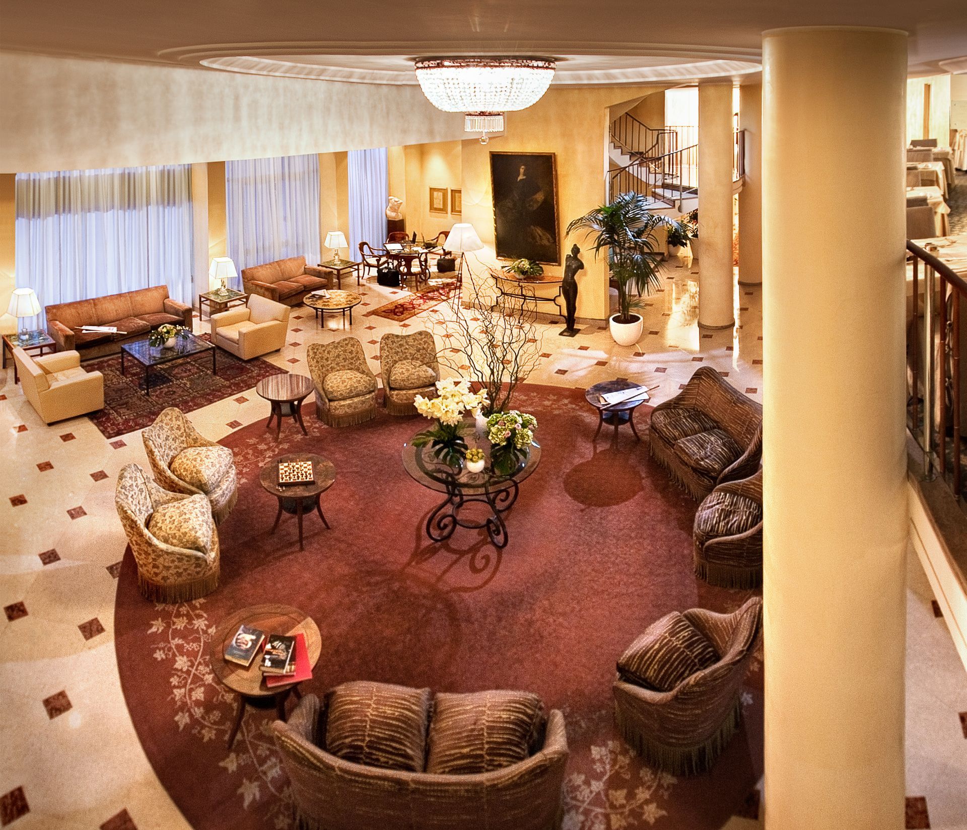 Vue plongeante sur le hall spacieux d'un hôtel, orné d'un tapis circulaire, de différents espaces de détente, d'un éclairage chaleureux et de colonnes.