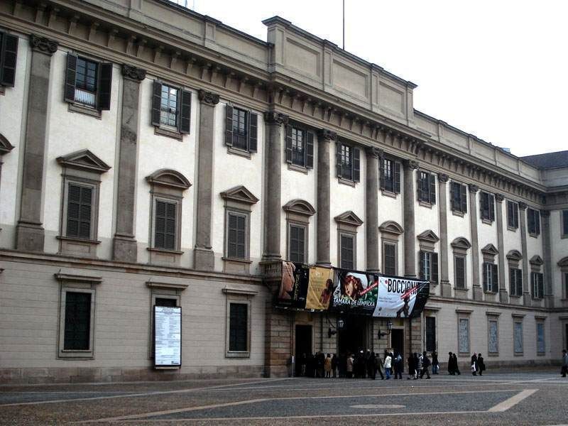 A long, neoclassical stone building with many windows and banners hanging over the entrance, with people gathered below.