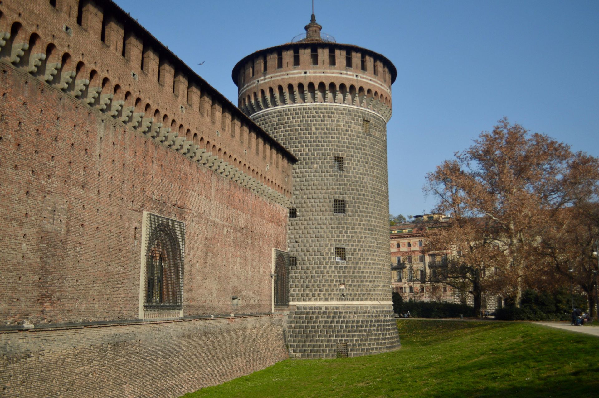 A brick fortress wall connects to a tall, cylindrical stone tower at Sforzesco Castle in Milan, surrounded by a grassy moat.