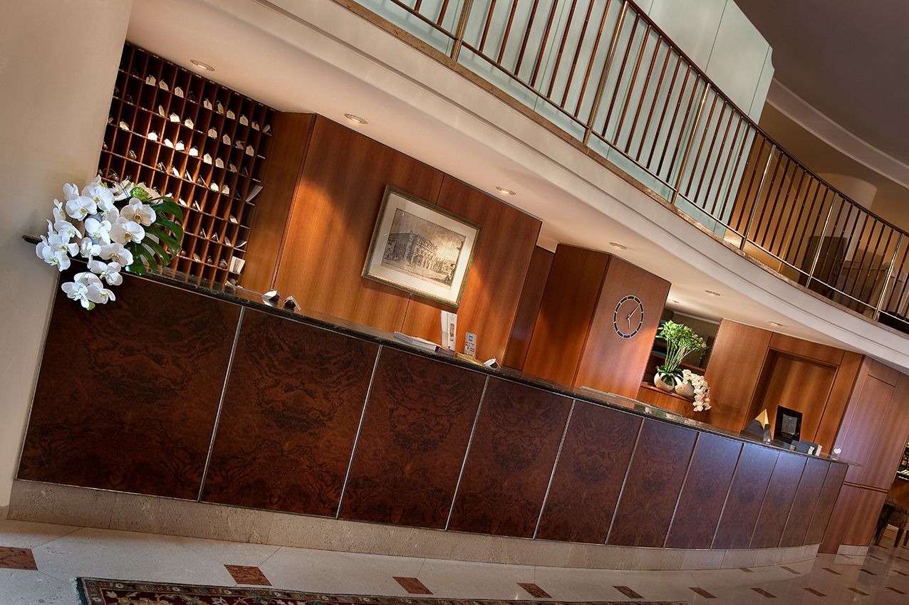 Стойка регистрации в холле отеля с темными деревянными панелями, изящными металлическими элементами и лестничным балконом на заднем плане.