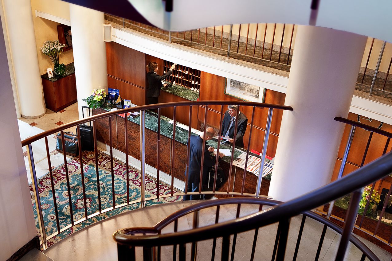 Кадр, снятый сверху, лобби отеля с деревянной стойкой регистрации, узорчатым ковром и изогнутой лестницей.