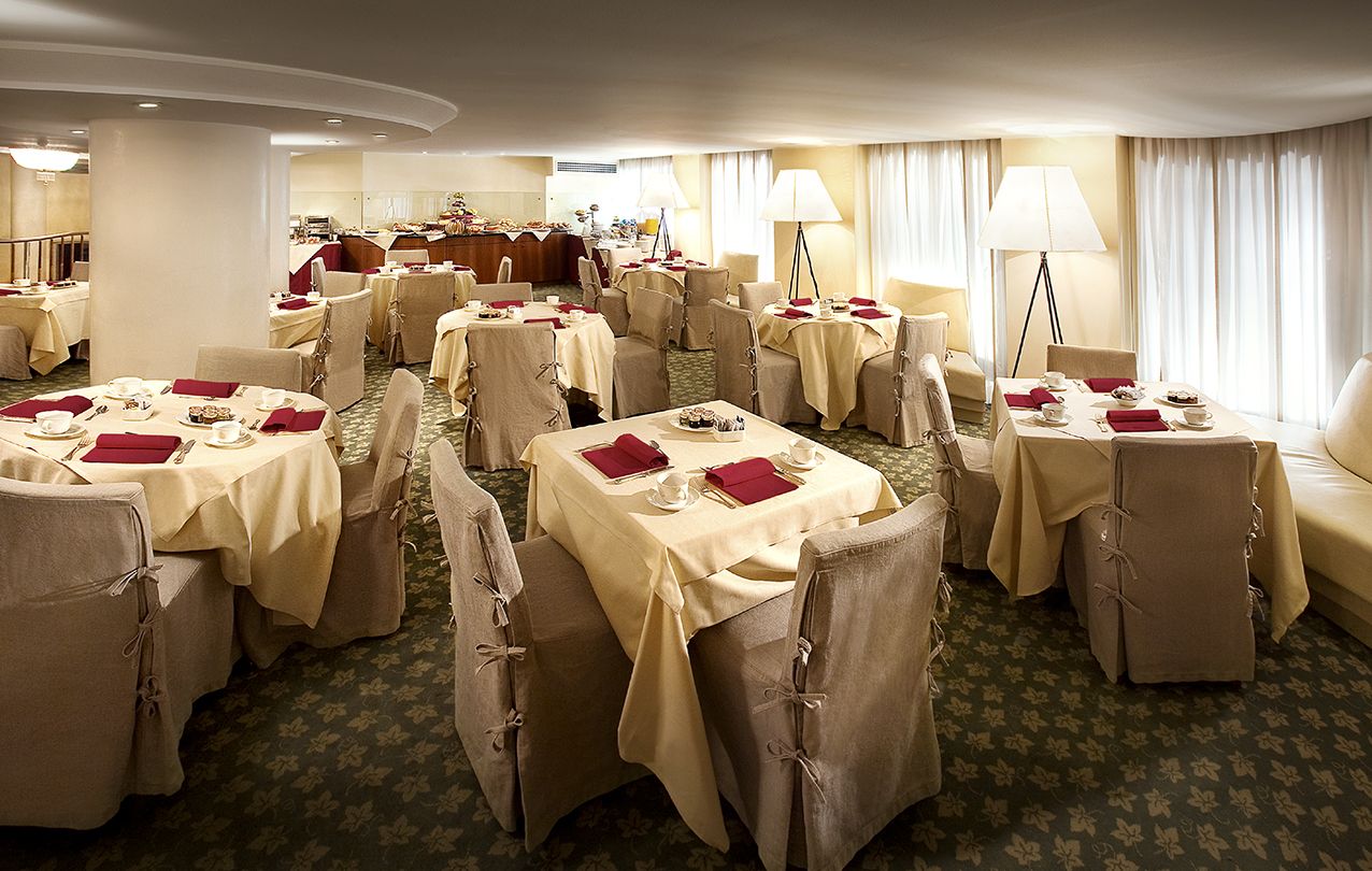 В столовой несколько столов с кремовыми скатертями, бордовыми салфетками и стульями в чехлах, расположенные на ковре с узором.