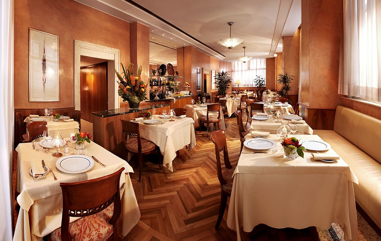 Элегантный, уютно освещенный обеденный зал со столами, сервированными для обслуживания, с паркетным полом «елочкой» и стенами нежно-бежевого цвета.
