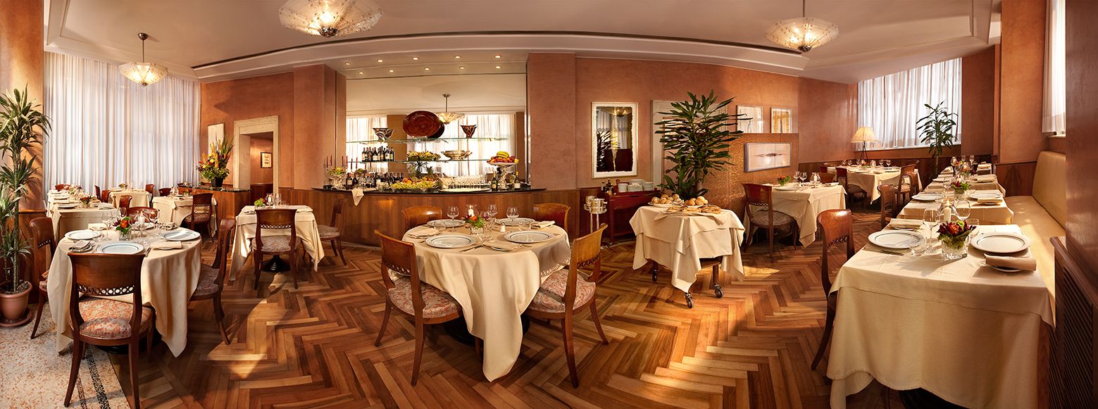 Ресторанный зал с множеством круглых и прямоугольных столов, покрытых белыми скатертями, на деревянном полу.