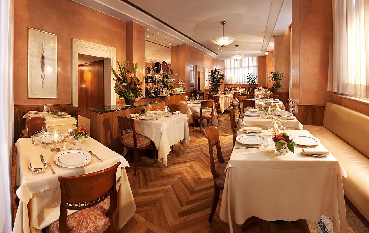 Элегантный обеденный зал ресторана с белыми скатертями на столах, деревянными стульями и стенами теплых оранжевых тонов.