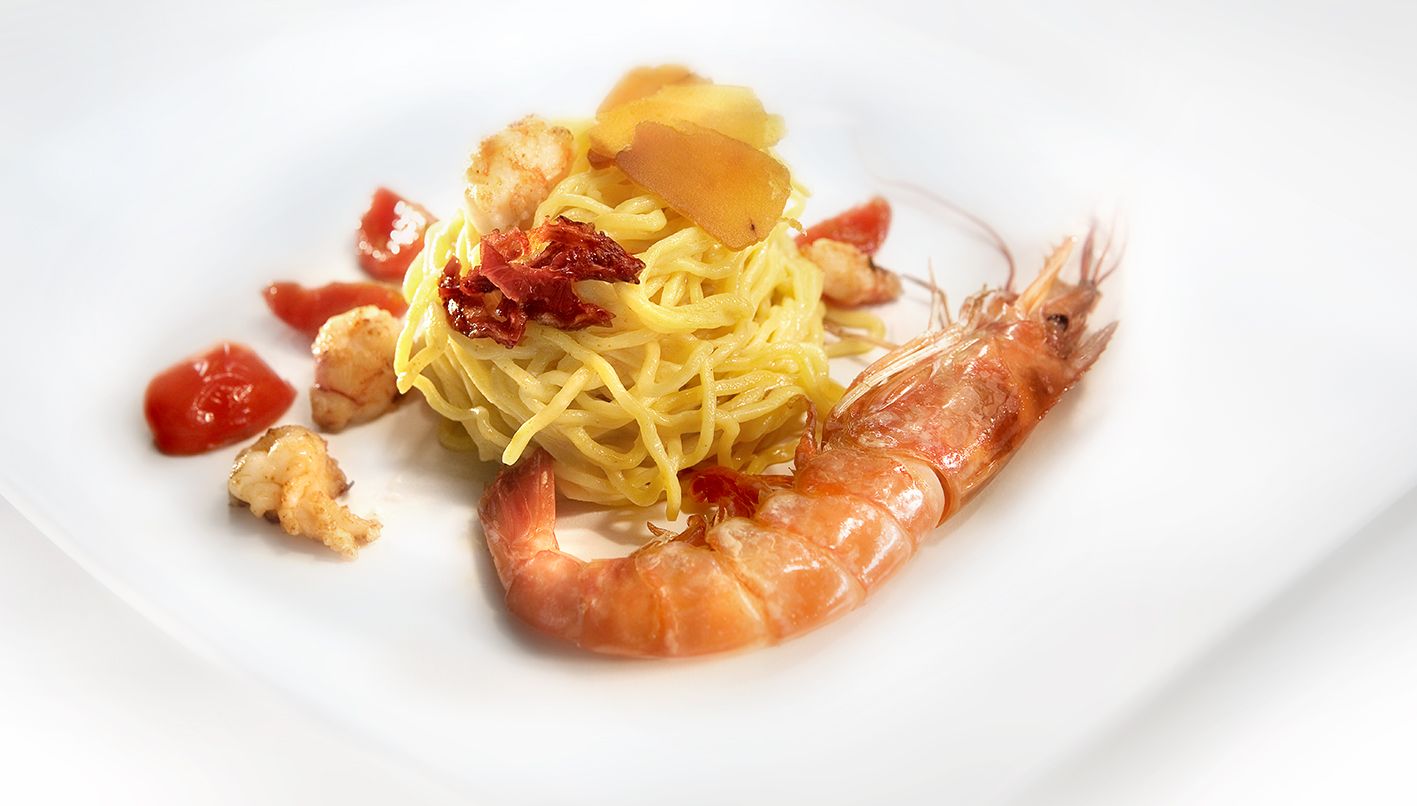Порция пасты, выложенной в форме гнезда, украшенная вялеными помидорами, в окружении креветок и жареных помидоров черри.