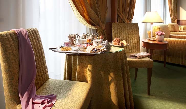 Небольшой круглый столик, накрытый для завтрака, стоит между двумя мягкими креслами в комнате с теплым освещением и драпированными шторами.